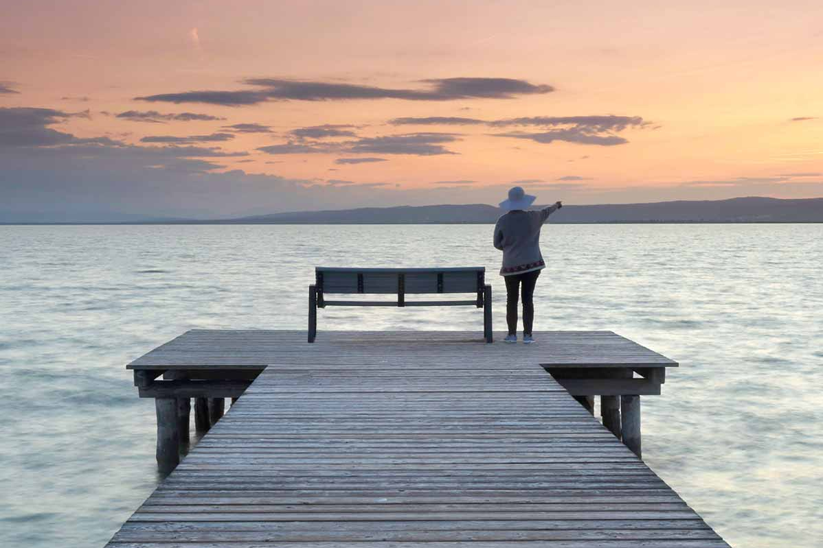 Betriebsvermögen in ETFs anlegen Eine Frau mit Hut steht auf einem Steg im See und zeigt auf eine Hügelkette